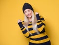 Excited beautiful young woman with closed eyes and clenched fists, model wearing woolen cap and sweater, isolated on yellow Royalty Free Stock Photo