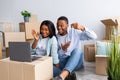 Excited black couple chatting online with friends and showing key of their house to camera, sitting among carton boxes Royalty Free Stock Photo