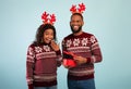 Excited black man giving wife Christmas gift, happy woman and her husband wearing deer horns, blue background