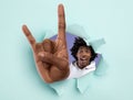 Excited black teenager showing huge rock gesture through hole in light blue ripped paper Royalty Free Stock Photo