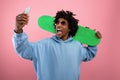 Excited black teenager with skateboard deck taking selfie on smartphone over pink studio background