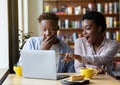 Excited black woman and her boyfriend pointing at laptop screen, shocked to see unexpected news in coffee shop