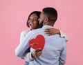 Excited black woman holding heart shaped gift box and hugging her boyfriend on pink studio background Royalty Free Stock Photo