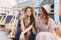 Excited blonde girl in transparent white dress looking at sister which posing holding wineglass. Outdoor portrait of two