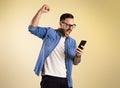 Excited businessman dressed in denim shirt screaming and raising hand while reading good news over mobile phone. Young man Royalty Free Stock Photo