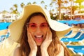 Excited cheerful girl with straw hat and sunglasses on the beach with umbrellas and deck chairs with palm trees on the background.