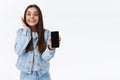 Excited, cheerful happy lively girl in denim jacket, jeans, holding smartphone, showing mobile phone screen as reacting