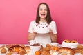 Excited cheerful woman wearing white T-shirt sitting at festive table with various desserts, isolated over pink background posing Royalty Free Stock Photo