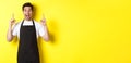 Excited coffee shop owner in black apron pointing fingers up, showing special offers, standing over yellow background