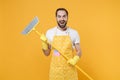 Excited crazy young man househusband in apron rubber gloves hold in hands broom while doing housework isolated on yellow