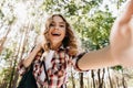 Excited curly girl making selfie on nature background. Winsome blonde woman in checkered shirt taki Royalty Free Stock Photo