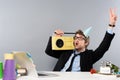 Excited drunk businessman sitting at workplace in party cap with vintage tape recorder on grey background. Royalty Free Stock Photo