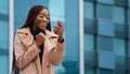 Excited enthusiastic surprised young woman winner standing outdoors reading good news on phone celebrating victory