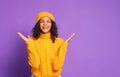 Excited ethnic female in warm yellow sweater and hat shouts with joy and gestures against purple background