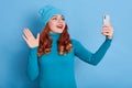 Excited female wearing blue jumper and cap, posing with raising palm, having video call or taking selfie, looks happy, wears red