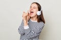 Excited festive woman with brown hair wearing striped shirt standing isolated over gray background listening to music on headphone
