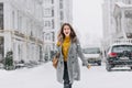 Excited girl in gray coat and ripped jeans walking down the road in snowy day. Trendy caucasian woman spending time