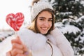 Excited girl in white attire enjoying winter vacation in forest and posing with candy. Smiling european woman in knitted