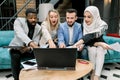 Excited group of diverse multiracial coworkers looking at the laptop screen with open mouth and smile, while sitting on