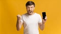 Excited guy holding cellphone shouting shaking fists, yellow background, mockup Royalty Free Stock Photo