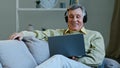 Excited happy aged man watching movie using laptop and headphones sitting lying on comfy couch smiling elderly Royalty Free Stock Photo