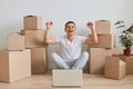 Excited happy smiling man wearing white T-shirt sitting on the floor near cardboard boxes with his pile, working on laptop, Royalty Free Stock Photo