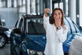 Excited with her new car. Holds keys in hand. Young woman in white official clothes stands in front of blue automobile