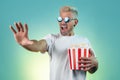 Excited hipster blonde man wearing stylish sunglasses and holding bucket with popcorn in studio in neon lights. Royalty Free Stock Photo