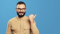 Excited hipster man points aside on empty space, isolated over blue wall