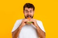 Excited hungry Caucasian man biting delicious cheeseburger against yellow backdrop
