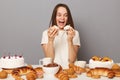 Excited hungry woman wearing white T-shirt isolated over gray background holding cakes expressing amazement breaking diet having