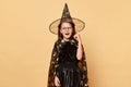 Excited inspired little girl wearing costume carnival cone hat and glasses isolated over beige background celebrating Halloween