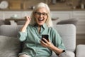 Excited joyful elderly woman in eyeglasses holding smartphone