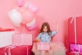 Excited joyful little girl in tulle skirt opening birthday present suround big colorful giftboxes on pink