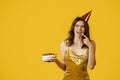 Excited lady in festive dress licking cream of birthday cake in her hands, wearing party hat and looking at free space