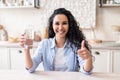 Excited latin woman holding glass with clean water and showing thumb up, sitting at table in kitchen interior Royalty Free Stock Photo