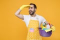 Excited man househusband in apron rubber gloves hold basin detergent bottles washing cleansers isolated on yellow