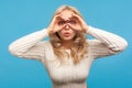 Excited nosy blond woman in white knitted sweater looking through holes in fingers like binoculars, spying, trying to find out Royalty Free Stock Photo