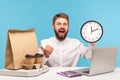 Excited positive bearded man office worker holding big wall clock pointing finger at paper bag, coffee cups and pizza box, Royalty Free Stock Photo