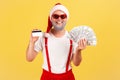 Excited positive gray bearded man in santa claus hat and sunglasses holding credit card and dollar banknotes looking at camera