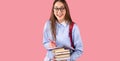 Excited school girl in uniform wearin glasses with backpack holding books and writing some notes in a diary on pink