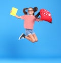 Excited schoolgirl in glasses jumping up from happiness and joy with backpack and exercise book in hands