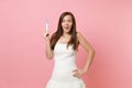 Excited shocked bride woman in white wedding dress holding pregnancy test on pastel pink background. Medical