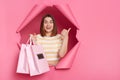 Excited surprised brunette woman wearing striped shirt looking through breakthrough of pink background, standing with shopping