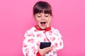 Excited surprised female child in casual style sweater holding smart phone in hands and keeping mouth opened, touching her cheek Royalty Free Stock Photo