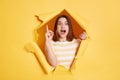 Excited surprised smart young adult woman stands in torn paper hole, having great idea, looking at camera with raised finger, Royalty Free Stock Photo