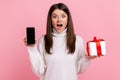 Excited surprised woman holding wrapped present box and cell phone with empty screen for promotion. Royalty Free Stock Photo