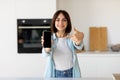 Excited woman holding smartphone with blank screen and pointing on it while standing in kitchen interior, copy space Royalty Free Stock Photo