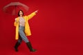 Excited woman in yellow waterproof raincoat and boots walking with umbrella and pointing at free space on red background Royalty Free Stock Photo