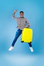 Excited young African American guy with yellow suitcase jumping and gesturing YES on blue studio background Royalty Free Stock Photo